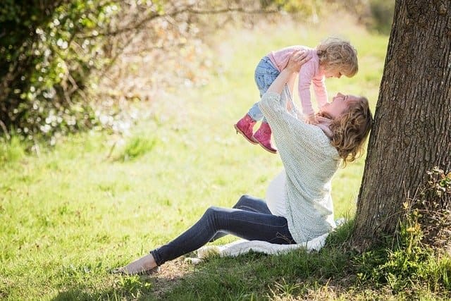 Photo d'une maman assise contre un arbre soulevant sa fille à bout de bras au dessus d'elle